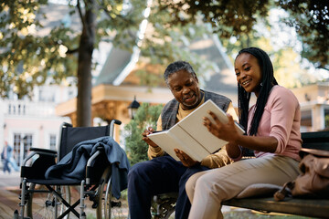 Happy black senior and his daughter looking at family photo album in park at nursing home.