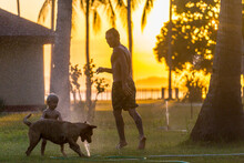 Father Playing With Dog And Baby Son Beside Splashing Garden Hose At Sunset, Kertasari, Sumbawa, Indonesia