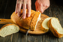 Close-up Of A Chef's Hands With A Knife Cutting Wheat Bread On A Kitchen Cutting Board. Bread On The Kitchen Table Or The Healthy Eating And Traditional Bakery Concept.