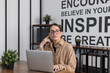 © Georgi - A young female office worker with glasses and a beige shirt sitting at her computer, pondering her work. Her desk features a wooden table, a notebook, a small plant, and various stationery items.