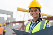 © Supachai - Asian woman engineer holding document smiling at construction site. Confident female Indian wearing protective helmet and vest working in factory making precast concrete wall for real estate housing.
