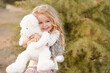 Smiling playful baby girl 4-5 year old holding teddy bear outdoors over green nature background. Childhood. Wearing stylish clothes.