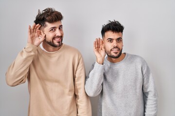 Wall Mural - Young homosexual couple standing over white background smiling with hand over ear listening an hearing to rumor or gossip. deafness concept.