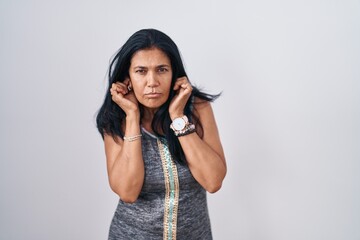 Sticker - Mature hispanic woman standing over white background covering ears with fingers with annoyed expression for the noise of loud music. deaf concept.