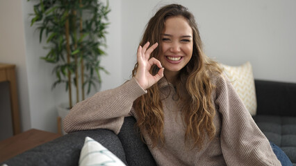 Young beautiful hispanic woman doing ok gesture sitting on sofa at home