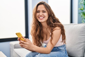 Young beautiful hispanic woman using smartphone sitting on sofa at home