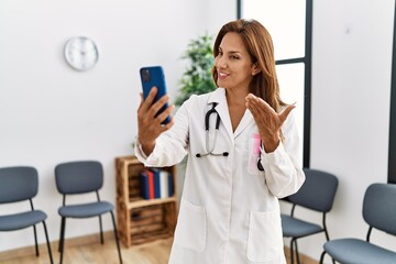 Canvas Print - Young latin woman wearing doctor uniform having video call at clinic waiting room