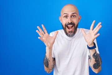 Poster - Hispanic man with tattoos standing over blue background celebrating crazy and amazed for success with arms raised and open eyes screaming excited. winner concept