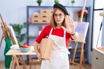 Sticker - Young redhead woman at art studio holding art case doing stop sing with palm of the hand. warning expression with negative and serious gesture on the face.