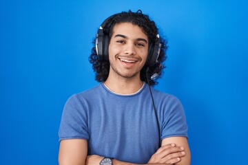 Wall Mural - Hispanic man with curly hair listening to music using headphones happy face smiling with crossed arms looking at the camera. positive person.