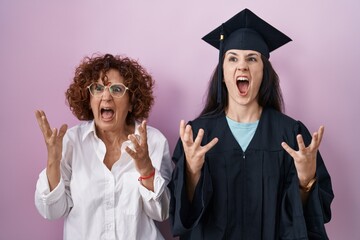 Wall Mural - Hispanic mother and daughter wearing graduation cap and ceremony robe crazy and mad shouting and yelling with aggressive expression and arms raised. frustration concept.