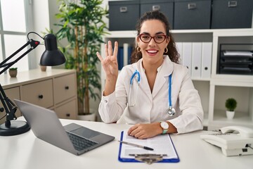 Sticker - Young hispanic woman wearing doctor uniform and stethoscope showing and pointing up with fingers number four while smiling confident and happy.