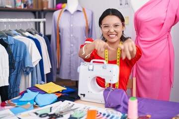 Wall Mural - Hispanic young woman dressmaker designer using sewing machine pointing to you and the camera with fingers, smiling positive and cheerful