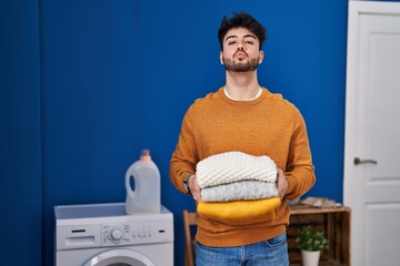 Wall Mural - Hispanic man with beard holding clean folded laundry looking at the camera blowing a kiss being lovely and sexy. love expression.