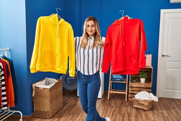 Poster - Young blonde woman holding clean laundry on hangers relaxed with serious expression on face. simple and natural looking at the camera.