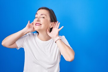 Canvas Print - Middle age hispanic woman standing over blue background trying to hear both hands on ear gesture, curious for gossip. hearing problem, deaf