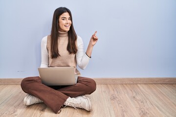 Wall Mural - Young brunette woman working using computer laptop sitting on the floor with a big smile on face, pointing with hand finger to the side looking at the camera.