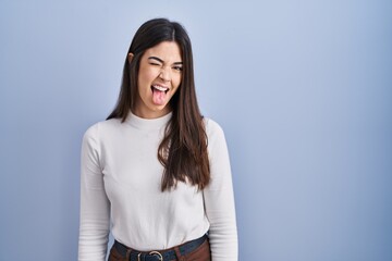 Sticker - Young brunette woman standing over blue background sticking tongue out happy with funny expression. emotion concept.