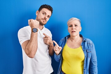 Poster - Young brazilian mother and son standing over blue background in hurry pointing to watch time, impatience, looking at the camera with relaxed expression