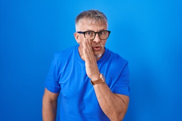 Wall Mural - Hispanic man with grey hair standing over blue background hand on mouth telling secret rumor, whispering malicious talk conversation