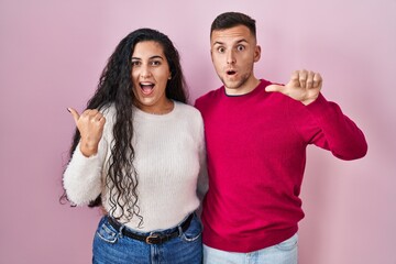 Wall Mural - Young hispanic couple standing over pink background surprised pointing with hand finger to the side, open mouth amazed expression.