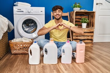 Sticker - Arab man with beard doing laundry sitting on the floor with detergent bottle smiling in love doing heart symbol shape with hands. romantic concept.