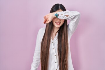 Wall Mural - Chinese young woman standing over pink background covering eyes with arm smiling cheerful and funny. blind concept.