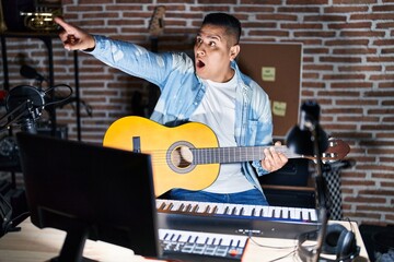 Wall Mural - Hispanic young man playing classic guitar at music studio pointing with finger surprised ahead, open mouth amazed expression, something on the front