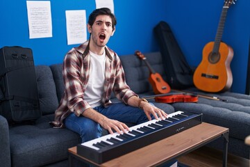 Poster - Young hispanic man playing piano at music studio angry and mad screaming frustrated and furious, shouting with anger. rage and aggressive concept.