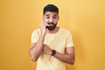 Canvas Print - Hispanic man with beard standing over yellow background looking stressed and nervous with hands on mouth biting nails. anxiety problem.