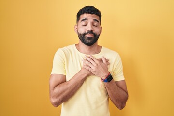 Sticker - Hispanic man with beard standing over yellow background smiling with hands on chest with closed eyes and grateful gesture on face. health concept.