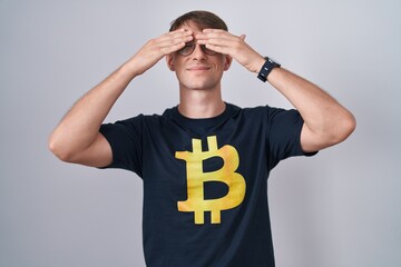 Sticker - Caucasian blond man wearing bitcoin t shirt covering eyes with hands smiling cheerful and funny. blind concept.