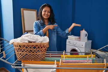 Wall Mural - Young asian woman hanging clothes at clothesline amazed and smiling to the camera while presenting with hand and pointing with finger.