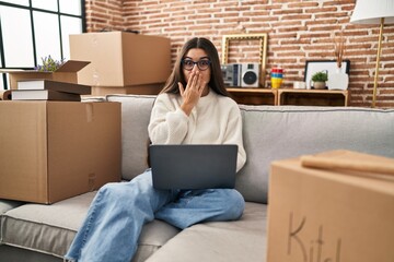 Poster - Young hispanic woman sitting on the sofa at new home using laptop smiling and looking at the camera pointing with two hands and fingers to the side.