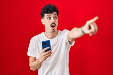 Canvas Print - Hispanic man using smartphone over red background pointing with finger surprised ahead, open mouth amazed expression, something on the front