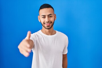 Poster - Young hispanic man standing over blue background smiling cheerful offering palm hand giving assistance and acceptance.