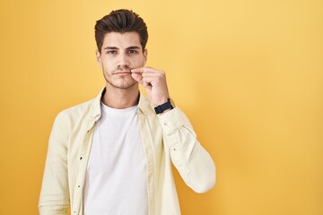 Sticker - Young hispanic man standing over yellow background mouth and lips shut as zip with fingers. secret and silent, taboo talking