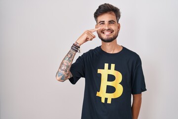 Poster - Young hispanic man with tattoos wearing bitcoin t shirt pointing with hand finger to face and nose, smiling cheerful. beauty concept