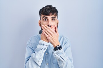 Wall Mural - Young hispanic man with beard standing over blue background shocked covering mouth with hands for mistake. secret concept.
