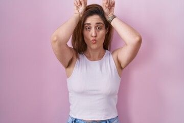 Wall Mural - Brunette woman standing over pink background doing funny gesture with finger over head as bull horns