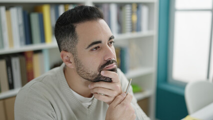 Sticker - Young hispanic man student sitting on table with doubt expression at library university