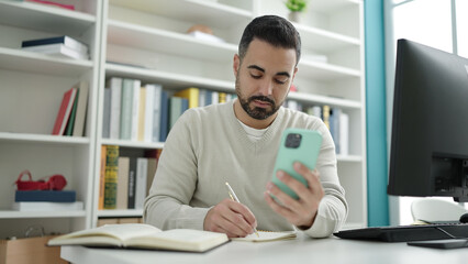 Sticker - Young hispanic man student using smartphone writing on notebook at library university