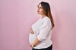 Pregnant woman standing over pink background looking to side, relax profile pose with natural face and confident smile.