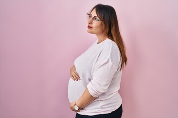 Poster - Pregnant woman standing over pink background looking to side, relax profile pose with natural face and confident smile.