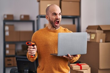 Canvas Print - Young bald man with beard working at small business ecommerce with laptop angry and mad screaming frustrated and furious, shouting with anger. rage and aggressive concept.