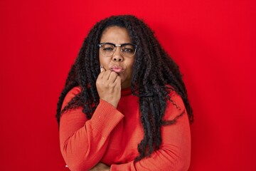 Sticker - Plus size hispanic woman standing over red background looking stressed and nervous with hands on mouth biting nails. anxiety problem.