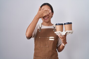 Wall Mural - Young hispanic woman wearing professional waitress apron holding coffee smiling and laughing with hand on face covering eyes for surprise. blind concept.