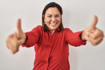 Wall Mural - Hispanic mature woman standing over white background approving doing positive gesture with hand, thumbs up smiling and happy for success. winner gesture.