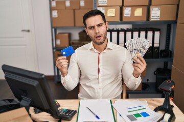 Sticker - Young hispanic man with beard working at small business ecommerce holding banknotes and credit card in shock face, looking skeptical and sarcastic, surprised with open mouth