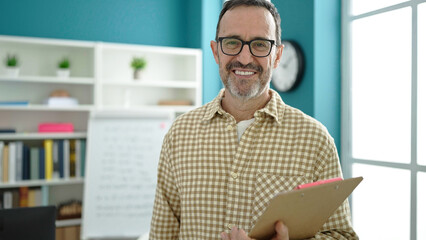 Middle age man teacher smiling confident holding clipboard at classroom